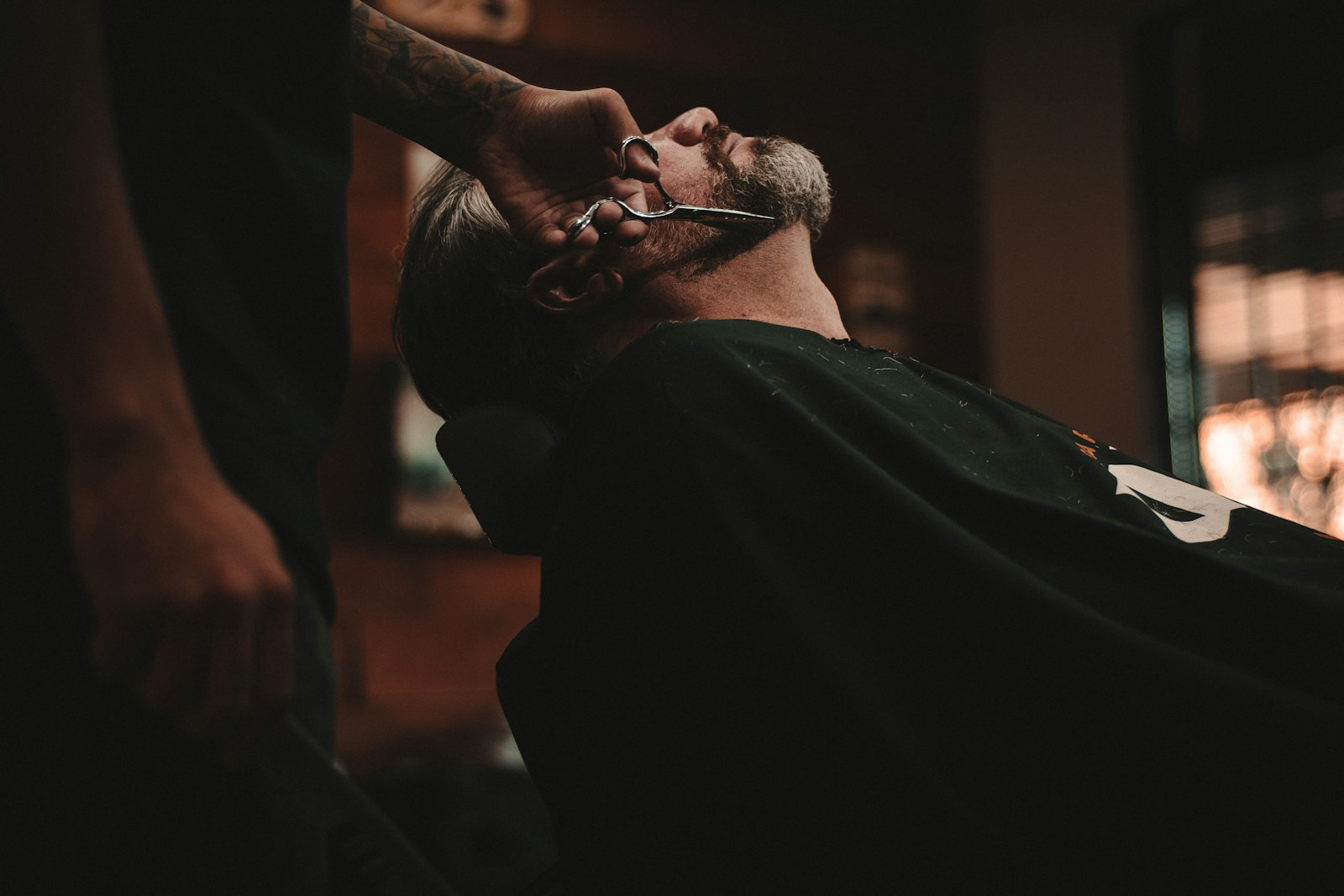 A barber giving a customer a beard trim with scissors in a warmly lit barbershop